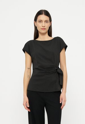 Woman with straight dark hair wears black pinstriped short-sleeve blouse with side tie, paired with black trousers, standing against white background.