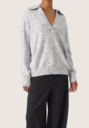 Personne portant un pull en maille gris clair ample avec un décolleté en V profond et un pantalon noir à jambes larges sur un fond clair.