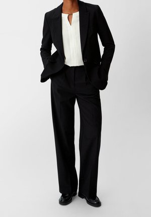 Frau trägt schwarzen Blazer mit passenden weit geschnittenen Hosen, weiße Bluse mit Schlüssellochausschnitt und schwarze glänzende Loafers, steht mit den Händen in den Taschen.