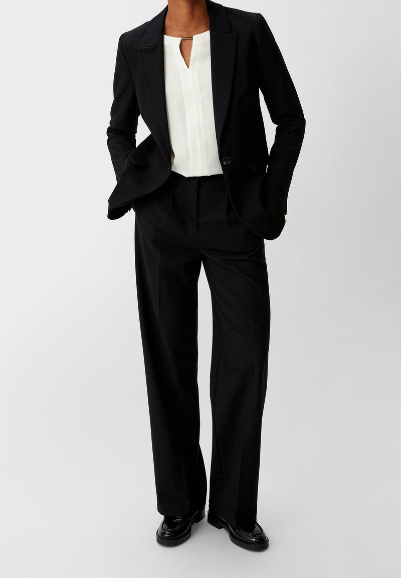 Femme portant un blazer noir assorti à un pantalon large, une blouse blanche avec un col en forme de goutte, et des mocassins noirs brillants, debout les mains dans les poches.