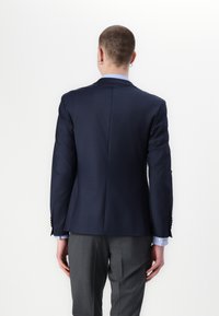 Marineblå blazer med strukturerte skuldre, enkel ventil og to knapper i front. Grå bukser med subtilt mønster og oppbrettede hem.