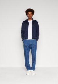 Marineblå zip-jakke med hvite striper, hvit T-skjorte, mørkeblå denimbukser og hvite sneakers. Avslappet passform, med synlig Lacoste-logo på jakken.