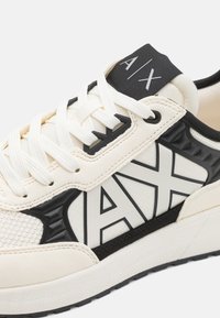 Valkoiset ja mustat tennarit, joissa on teksturoitu kangas, rohkea logo ja kontrastivärit. Kumipohja tarjoaa vakautta ja pitoa.