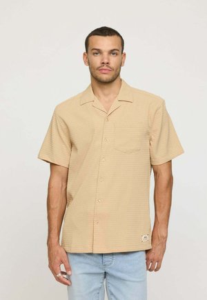 Chemise beige à manches courtes à boutons, avec un motif texturé, une poche poitrine unique et un ourlet droit. Portée avec un jean bleu clair.