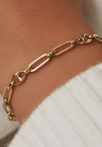 Bracelet en chaîne dorée avec des maillons ovales et ronds ; surface lisse ; porté au poignet ; le fond présente un tissu blanc doux et texturé.