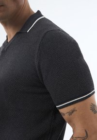 Camisa polo negra de manga corta con tejido texturizado, acentos en blanco y marino en el cuello y los puños de las mangas.
