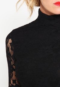 Top negro de encaje con cuello alto que presenta mangas largas, un intrincado patrón floral y detalles transparentes en las mangas. Tejido texturizado.
