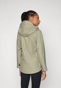 Jachetă impermeabilă de un verde măslinie deschis, cu glugă, mâneci lungi și un tiv curbat. Are un design elegant și un stil minimalist.