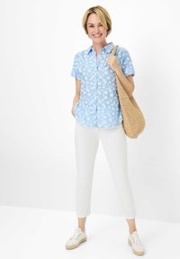 Korte mouwen blauwe overhemd met wit bloemenpatroon, gecombineerd met witte enkellange broek en een beige gevlochten tas. Witte sneakers maken de look compleet.