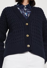 Marinblå stickad kofta med strukturerat mönster, djup V-hals och fem gyllene knappar, med ribbade ärmslut och fåll.