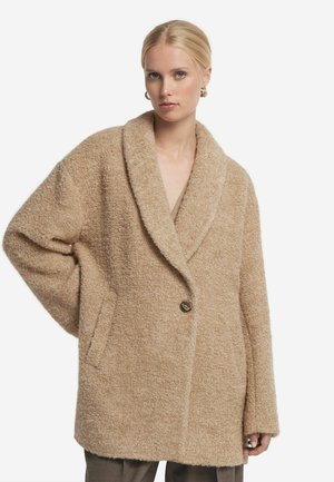 Manteau oversized beige en tissu texturé et duveteux, col à revers large, fermeture à un bouton et poches latérales. Silhouette simple et décontractée.