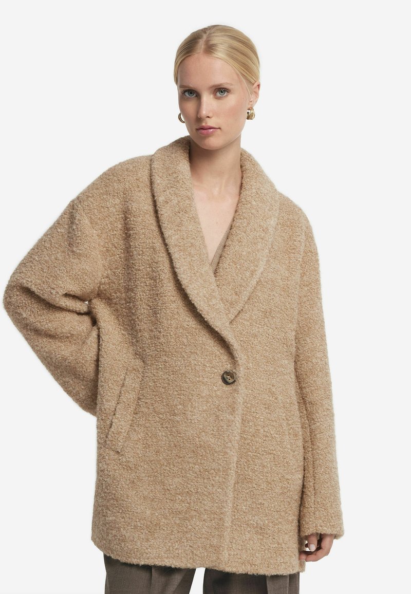 Beige överdimensionerad kappa i ett strukturerat, mjukt tyg, med stor slagkrage, ensamt knappstängning och sidofickor. Enkel, avslappnad silhuett.