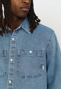Denim overhemd in lichtblauw. Voorzien van knoopzakken, een klassieke kraag en een label aan de linkerkant. Materiaaltextuur is soepel en casual.