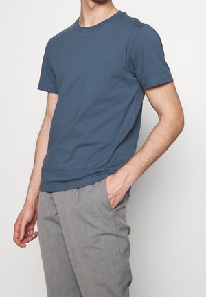 Homme portant un t-shirt bleu uni à manches courtes et un pantalon gris clair, avec une main dans la poche, sur un fond blanc.