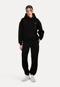 Felpa nera in pile con tasca frontale e logo, abbinata a pantaloni sportivi neri. L'outfit ha una vestibilità comoda, una consistenza morbida e polsini. Sneakers bianche.