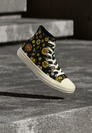 High-top sneaker med hvit såle og lisser, svart stoff dekket med fargerik blomsterbroderi, posisjonert i luften over betongtrinn.