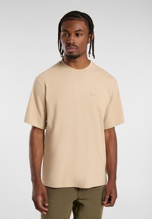 Beige Baumwoll-T-Shirt mit kurzen Ärmeln, Rundhalsausschnitt und einem kleinen Markenpatch auf der linken Brust. Glatte Textur und lässiger Schnitt.