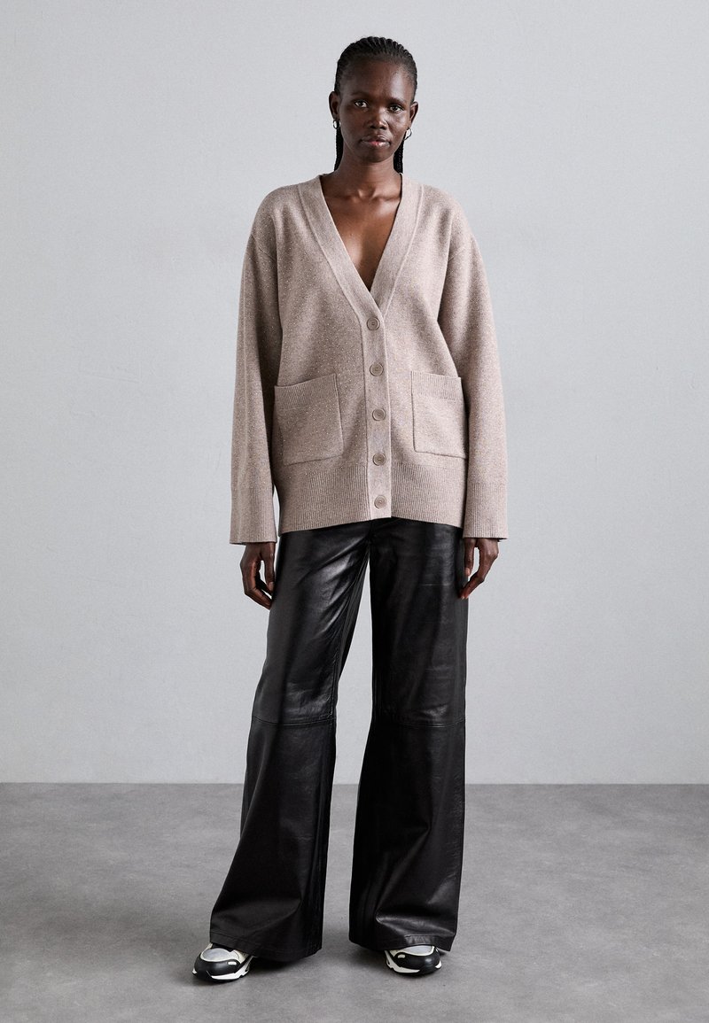 Beige Strickcardigan mit V-Ausschnitt, vier Knöpfen und zwei vorderen Taschen. Kombiniert mit schwarzen, weit geschnittenen Lederhosen und sportlichen Turnschuhen.