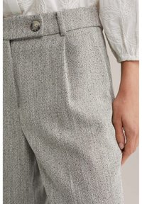 Grijze broek met een visgraatpatroon en een getextureerde stof, voorzien van een knoopsluiting en zijzakken, afgebeeld met een gedeeltelijke hand.