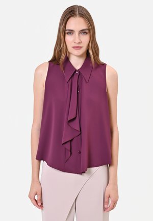 Donna con capelli lisci castano chiaro indossa una camicetta viola senza maniche con volant sul davanti e gonna beige chiaro a vita alta su sfondo bianco.