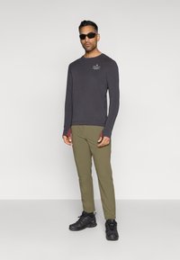 Σκούρος γκρι μπλουζάκι με μακριά μανίκια και διακριτικό γραφικό, συνδυασμένο με ελαιόχρωμες στενές παντελόνες και μαύρα αθλητικά παπούτσια με υφή.