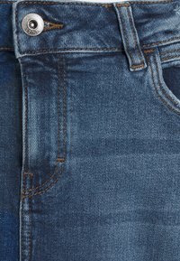Pantalones vaqueros de mezclilla azul con cierre de botón plateado, costuras naranjas que contrastan y textura ligeramente desvaída cerca de los bolsillos.