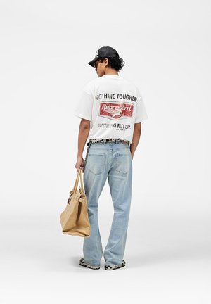 Hombre vistiendo camiseta blanca con estampado, jeans azul claro, gorra negra, cinturón y zapatos de leopardo, sosteniendo una bolsa grande beige, de espaldas sobre un fondo blanco.
