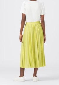 Personne portant un haut blanc à manches courtes, une jupe midi plissée vert lime et des baskets blanches, se tenant de dos contre un fond uni.