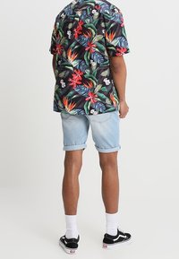 Camisa tropical de manga corta negra con patrones florales coloridos, combinada con pantalones cortos de mezclilla azul claro enrollados y zapatillas negras.