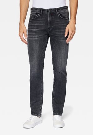 Mand iført slim-fit falmede sorte jeans og hvide sneakers, stående med armene afslappet ved siderne mod en hvid baggrund.