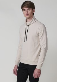 Giovane uomo che indossa una camicia beige a maniche lunghe con lacci neri e pantaloni neri, in piedi davanti a uno sfondo grigio chiaro.