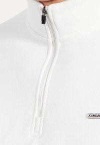 Sudadera blanca con cuello alto y media cremallera. Presenta una textura ligera y una pequeña etiqueta de logo plateado en el lado izquierdo.