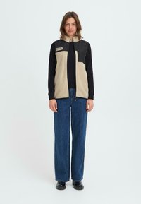 Beige Fleeceweste mit schwarzem Einsatz, Reißverschluss und Tasche. Getragen über einem schwarzen Langarmshirt, kombiniert mit blauen weit geschnittenen Jeans und schwarzen Schuhen.