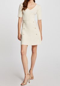 Knit-creme Kleid mit V-Ausschnitt, kurzen Ärmeln, figurbetontem Oberteil, ausgestellt Rock und goldenen Knopf-Akzenten entlang der Vorderseite.