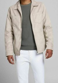 Beige mockaskinnjacka med dragkedja, med krage, en bröstficka och två sidofickor, över en grön stickad tröja.