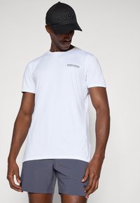 Λευκή κοντομάνικη αθλητική μπλούζα με μαύρο λογότυπο στο στήθος, συνδυασμένη με σκούρες γκρι βερμούδες με υφασμάτινο σχέδιο και λογότυπο. Μαύρο καπέλο με ανάγλυφα γράμματα.