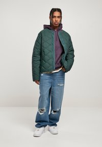 Chaqueta verde acolchada, sudadera morada con capucha, jeans claros de mezclilla desgastados y zapatillas blancas. La chaqueta tiene un cuello redondo y un cierre de botones a presión.