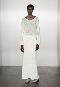 Abito lungo bianco in maglia con vestibilità morbida, dotato di scollo a barca e maniche lunghe. La modella indossa scarpe grigie e lo sfondo è neutro.