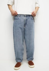 Persona vestida con pantalones vaqueros de denim ligeros y sueltos, sudadera blanca y zapatos tipo mocasín marrones, de pie contra un fondo blanco liso.