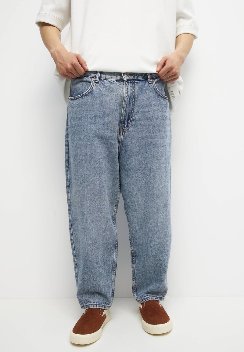 Persona vestida con pantalones vaqueros de denim ligeros y sueltos, sudadera blanca y zapatos tipo mocasín marrones, de pie contra un fondo blanco liso.