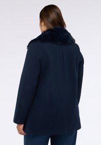 Cappotto blu navy con collo in pelliccia sintetica, silhouette dritta, maniche lunghe e texture liscia. Nessun hardware o motivo visibile.