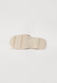Semelle de chaussure beige avec deux crêtes horizontales, motif de bande de roulement visible et légère courbure sur fond clair uni.