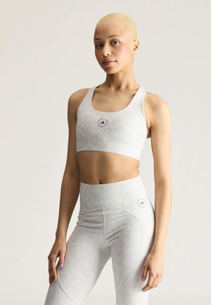 Jonge vrouw met kort blond haar, gekleed in een bijpassende lichtgrijze sportbeha en legging met hoge taille en subtiele patronen en logo's.