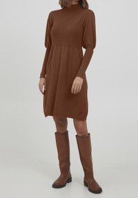 Langemouw, hoge hals bruine gebreide jurk met een gegatherde taille en pofmouwen, gedragen met kniehoog bruine leren laarzen.