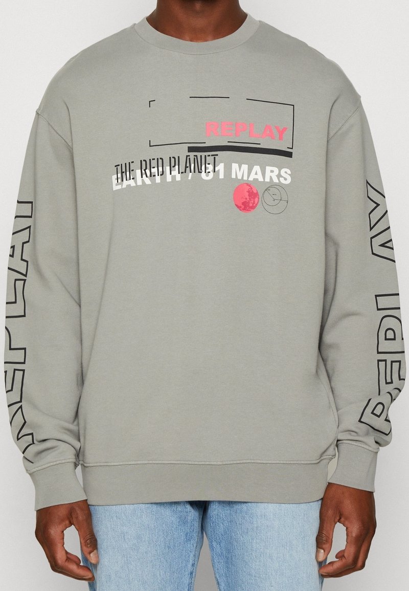 Sweat-shirt gris clair avec "REPLAY" et des textes et graphiques sur le thème de l'espace, porté par une personne à la peau de tonalité moyenne et un jean bleu clair.