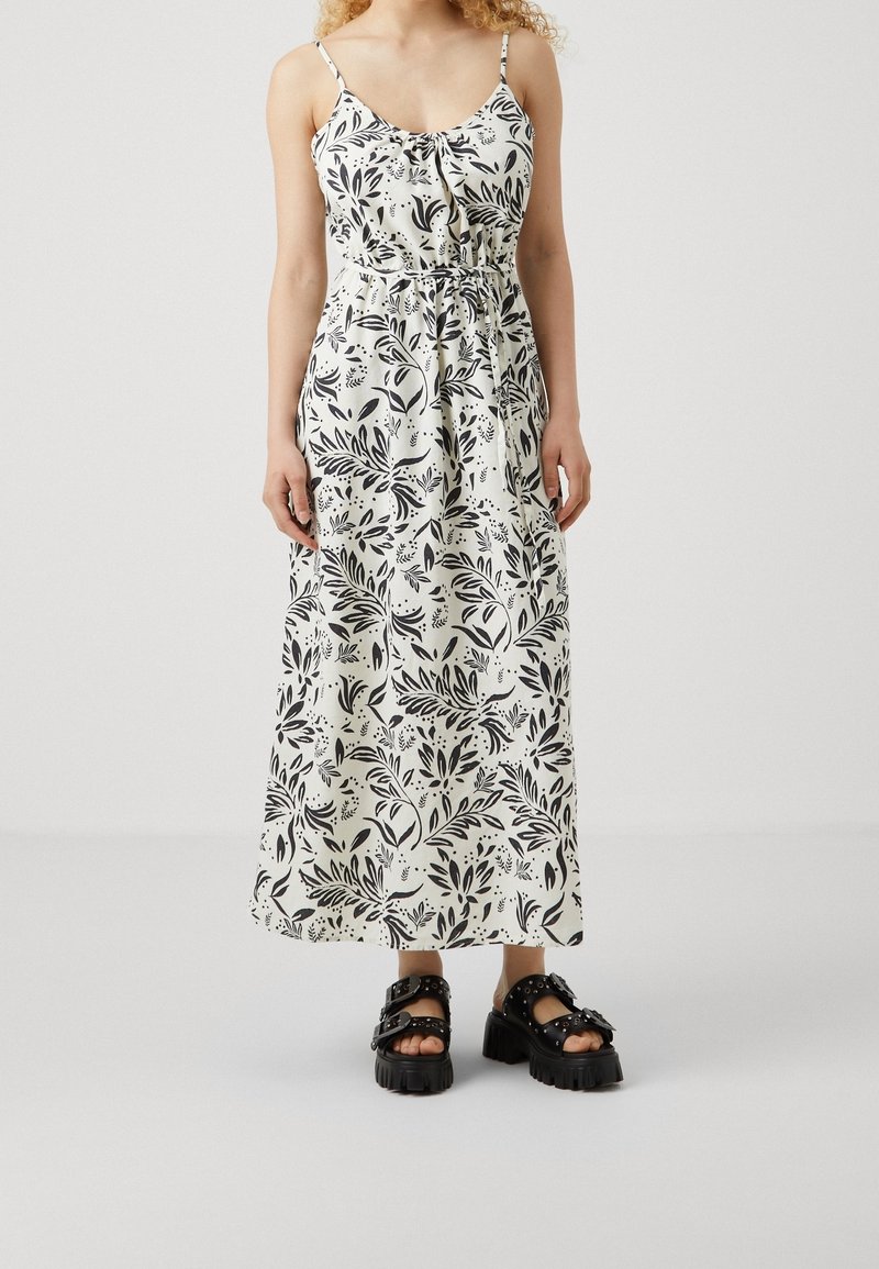 Femme portant une robe maxi blanche sans manches avec des motifs de feuilles noires et des sandales noires épaisses à boucles.