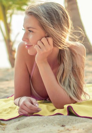 A person with long, light brown hair, wearing a pink swimsuit, rests on a yellow towel with pink edges on a sandy beach.