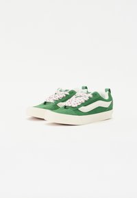 Grønn semsket sneakers med hvite detaljer, med et unikt rosa blomsterdesign på snørene og en tykk hvit gummisåle.