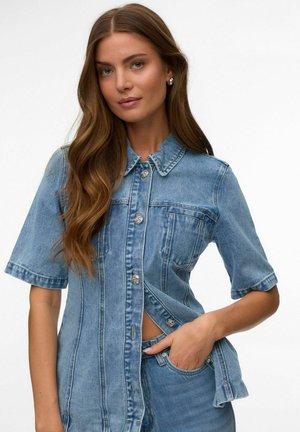 Jeune femme aux longs cheveux bruns portant une chemise en jean à manches courtes boutonnée et un jean assorti, une main dans la poche.