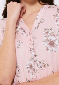 Blůza s květinovým vzorem ve světle růžové látce. Má V-výstřih, detail s knoflíkem a krátké rukávy s jemně zlatými květinovými akcenty.